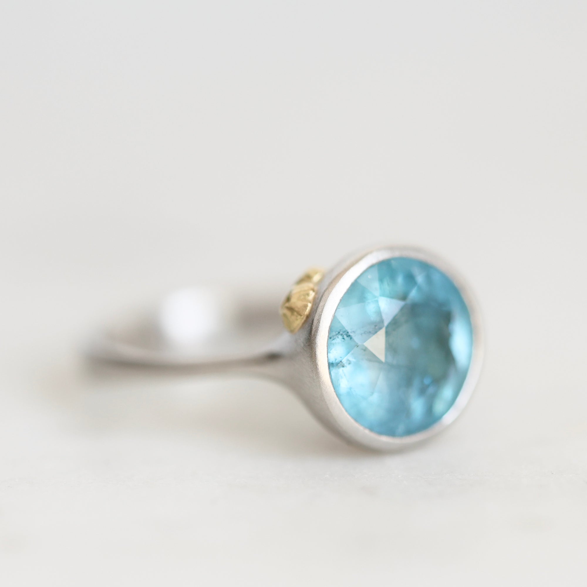 A silver ring featuring a round faceted light blue gemstone, with a small gold accent on the band, photographed against a soft, light gray background.