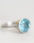 A silver ring featuring a round faceted light blue gemstone, with a small gold accent on the band, photographed against a soft, light gray background.