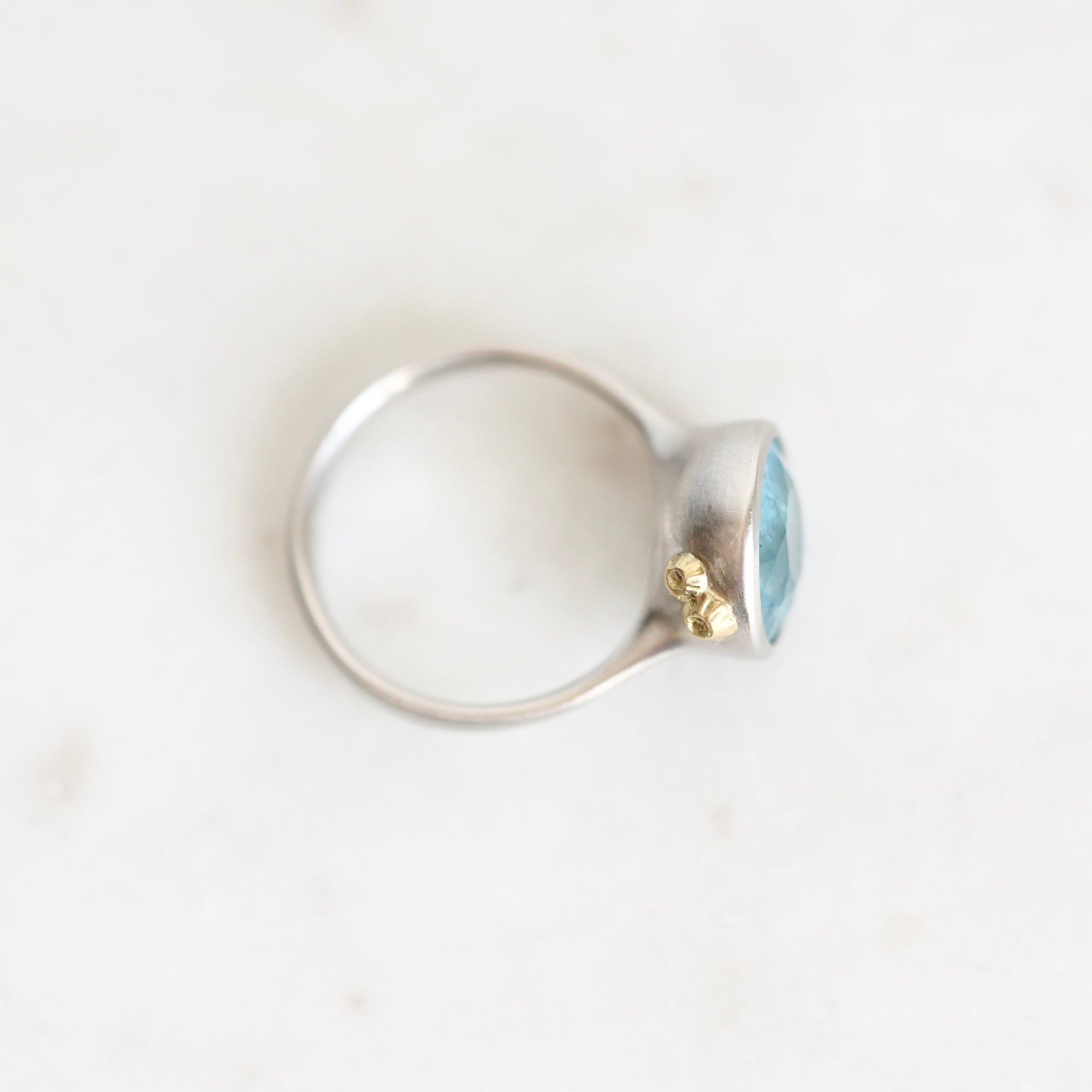A silver ring with a small gold accent and a light blue oval gemstone, photographed on a light off-white marble surface.