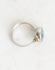 A silver ring with a small gold accent and a light blue oval gemstone, photographed on a light off-white marble surface.