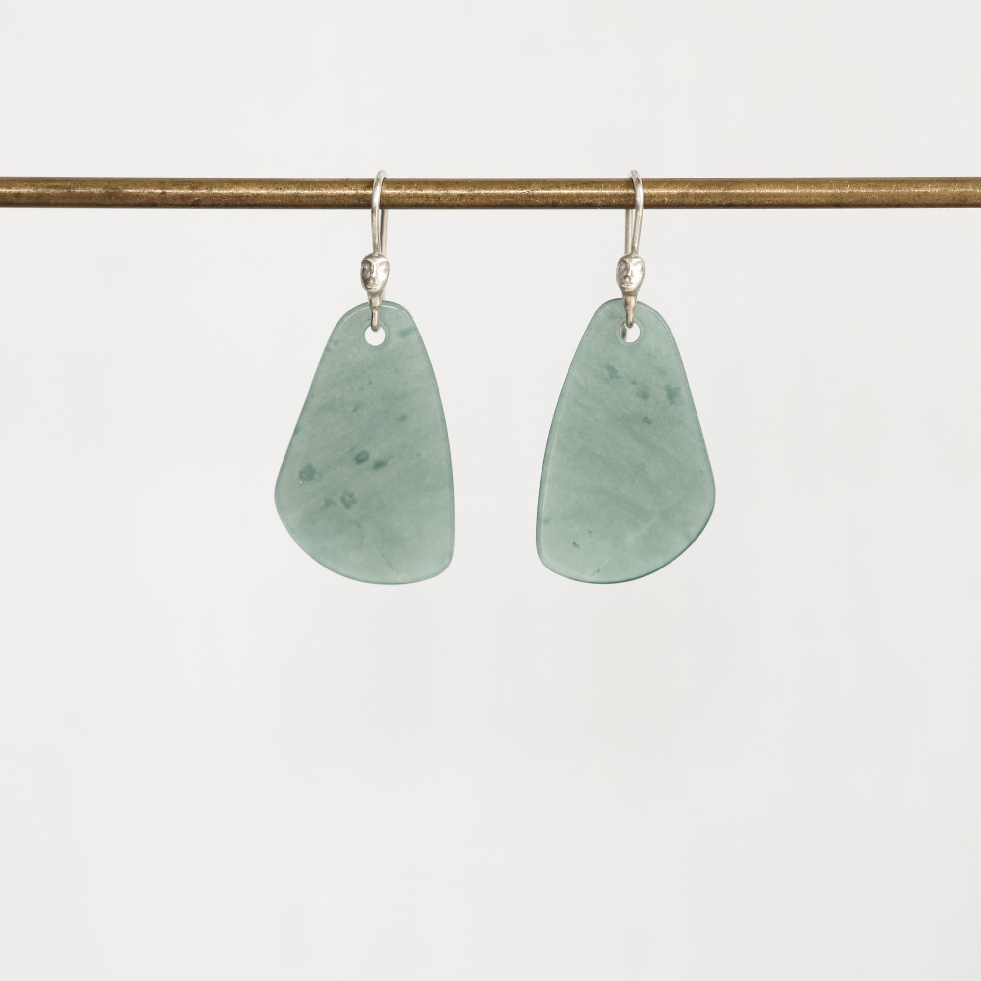 A pair of teardrop-shaped light green stone pendants on silver fishhook ear wires, hanging from a horizontal brass rod against a plain white background.