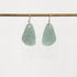 A pair of teardrop-shaped light green stone pendants on silver fishhook ear wires, hanging from a horizontal brass rod against a plain white background.
