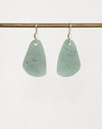A pair of teardrop-shaped light green stone pendants on silver fishhook ear wires, hanging from a horizontal brass rod against a plain white background.