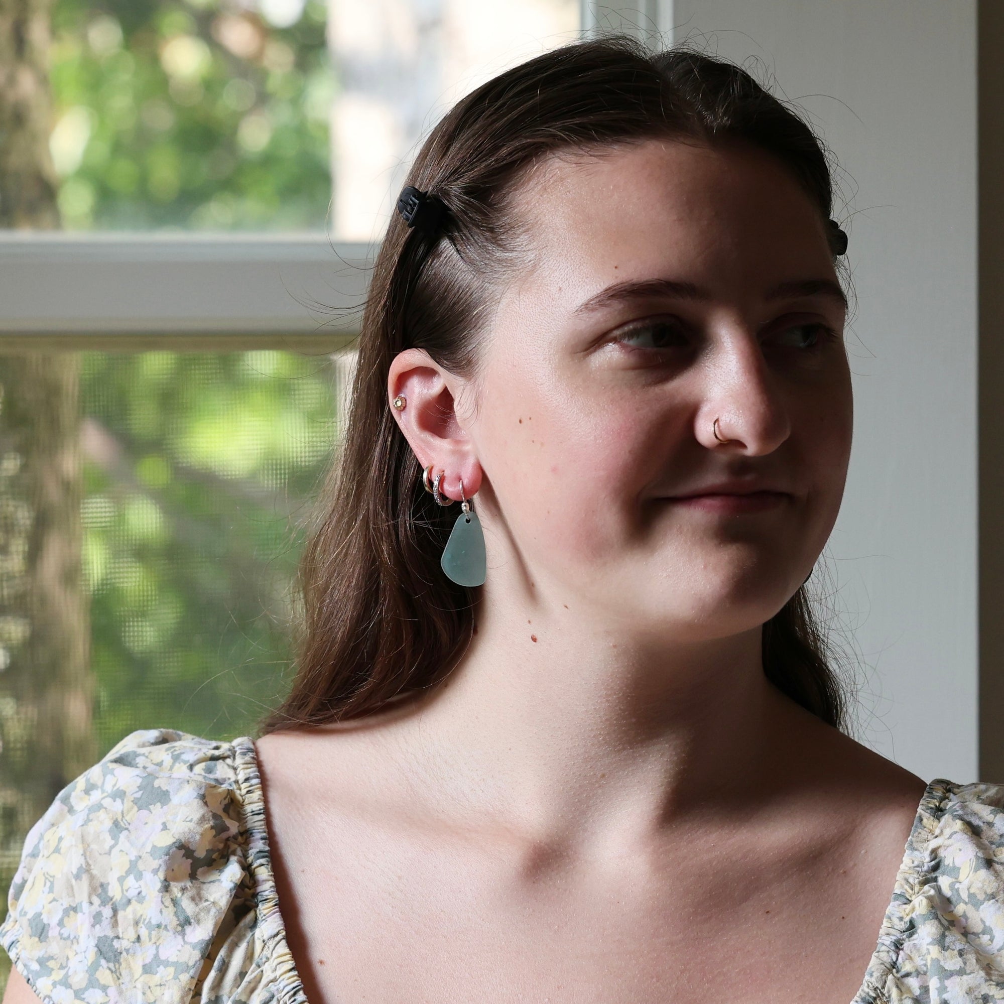 A close-up portrait of a woman with brown hair, wearing a floral puff-sleeve top and a light blue teardrop earring, standing in front of a window.