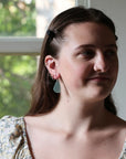 A close-up portrait of a woman with brown hair, wearing a floral puff-sleeve top and a light blue teardrop earring, standing in front of a window.