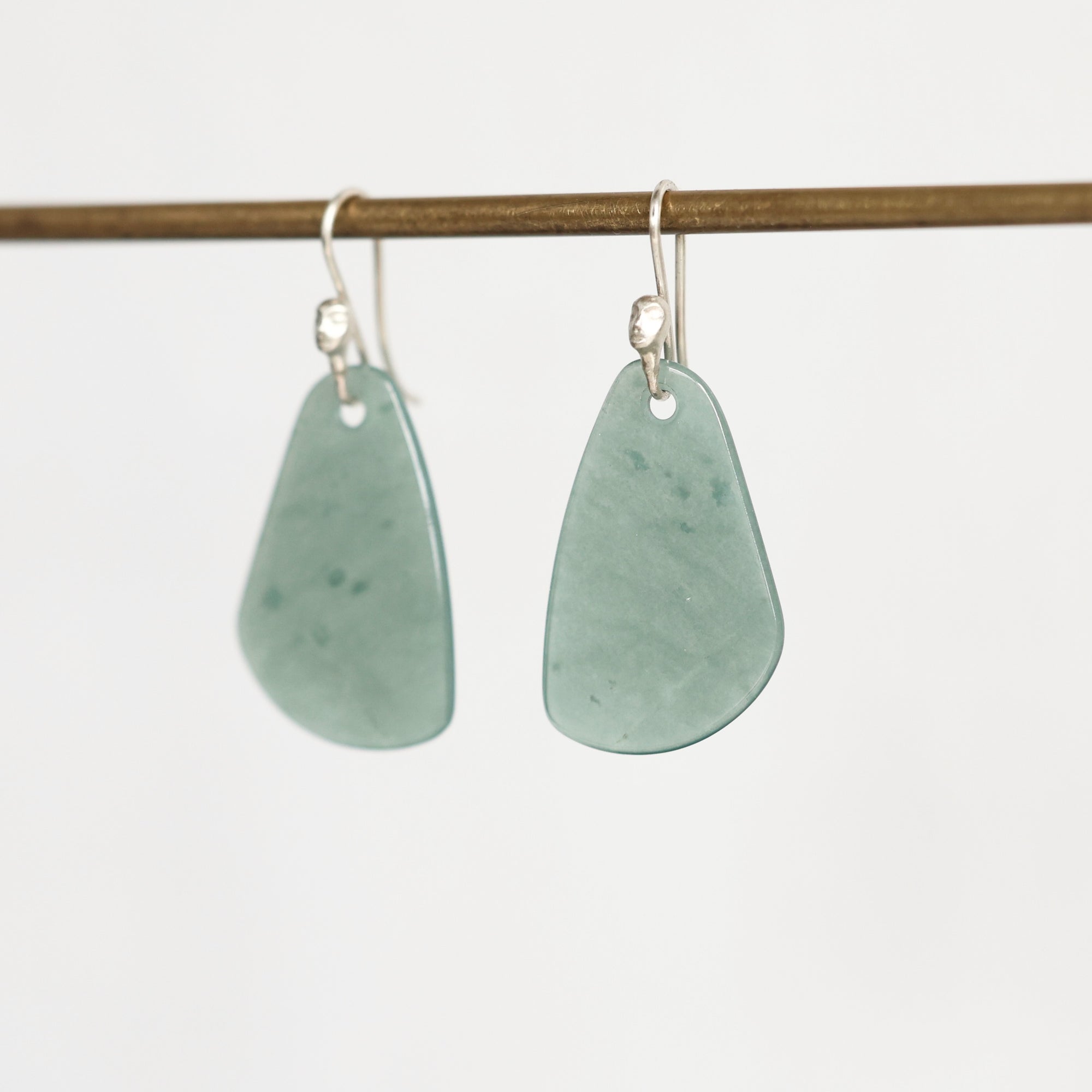 A pair of silver-toned dangle earrings with irregular, organic-shaped light green stone pendants, hanging from a thin horizontal wooden rod against a plain white background.