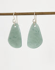 A pair of silver-toned dangle earrings with irregular, organic-shaped light green stone pendants, hanging from a thin horizontal wooden rod against a plain white background.