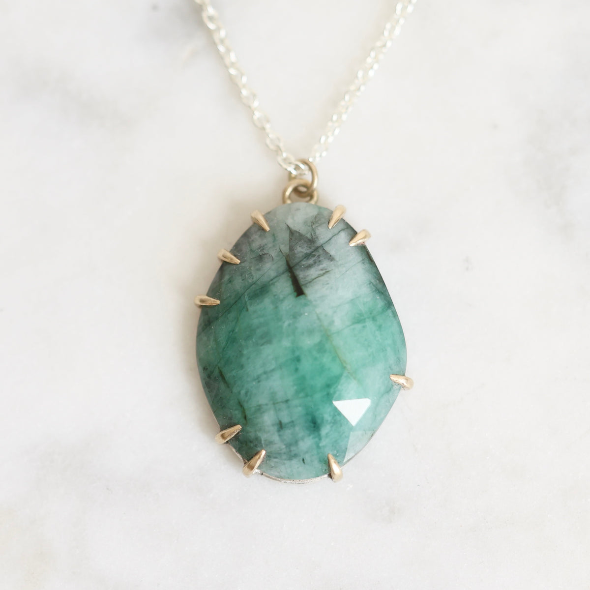 A teardrop-shaped green gemstone pendant with gold spiky accents around the edges, hanging from a delicate gold chain on a light marble background.