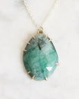 A teardrop-shaped green gemstone pendant with gold spiky accents around the edges, hanging from a delicate gold chain on a light marble background.