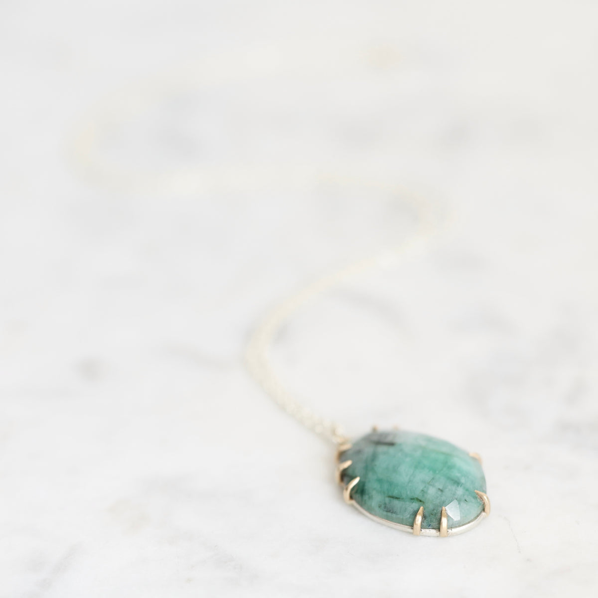 A green stone pendant necklace with a gold-toned bezel setting and delicate chain, resting on a light marble surface.