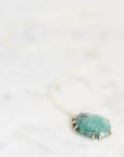 A green stone pendant necklace with a gold-toned bezel setting and delicate chain, resting on a light marble surface.