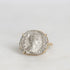 A gold ring featuring an antique silver coin with an embossed profile portrait, set against a plain light gray background.