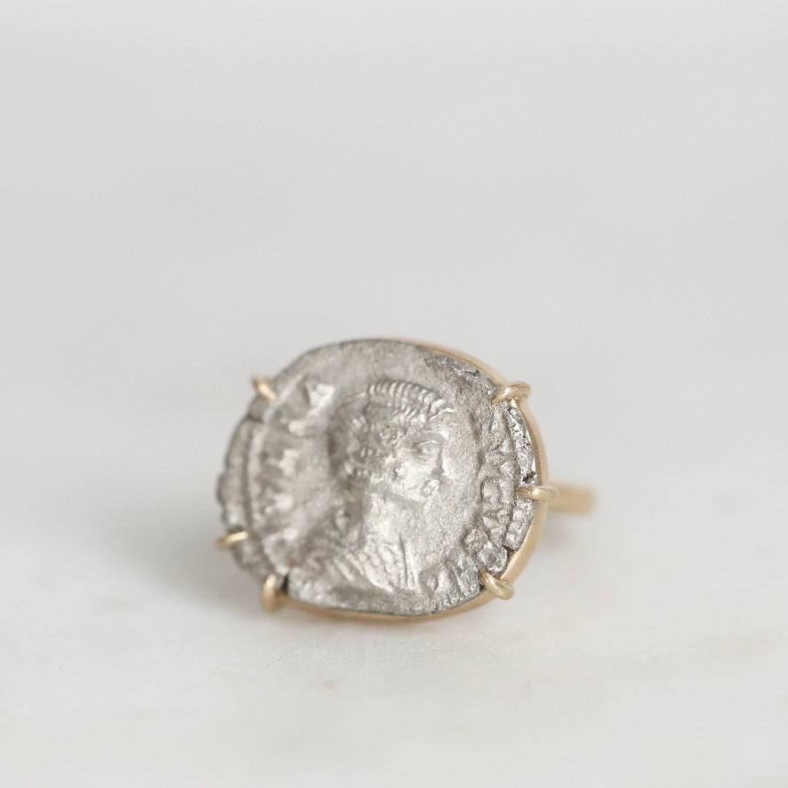 A gold ring featuring an antique silver coin with an embossed profile portrait, set against a plain light gray background.