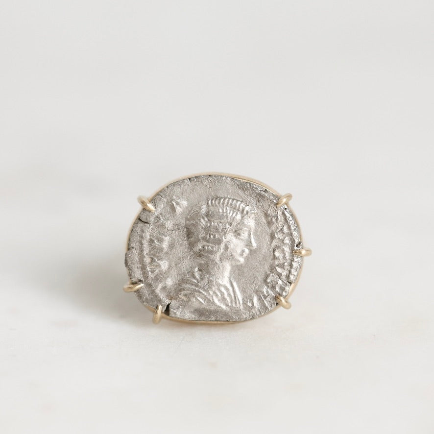 A silver-toned ring featuring an antique-style coin with a profile portrait, set in a small gold-toned prong setting against a light gray background.