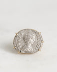 A silver-toned ring featuring an antique-style coin with a profile portrait, set in a small gold-toned prong setting against a light gray background.