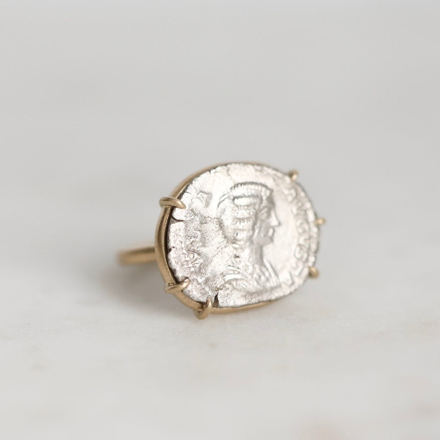 A gold ring featuring an antique silver coin with a profile portrait, set against a plain light gray background.