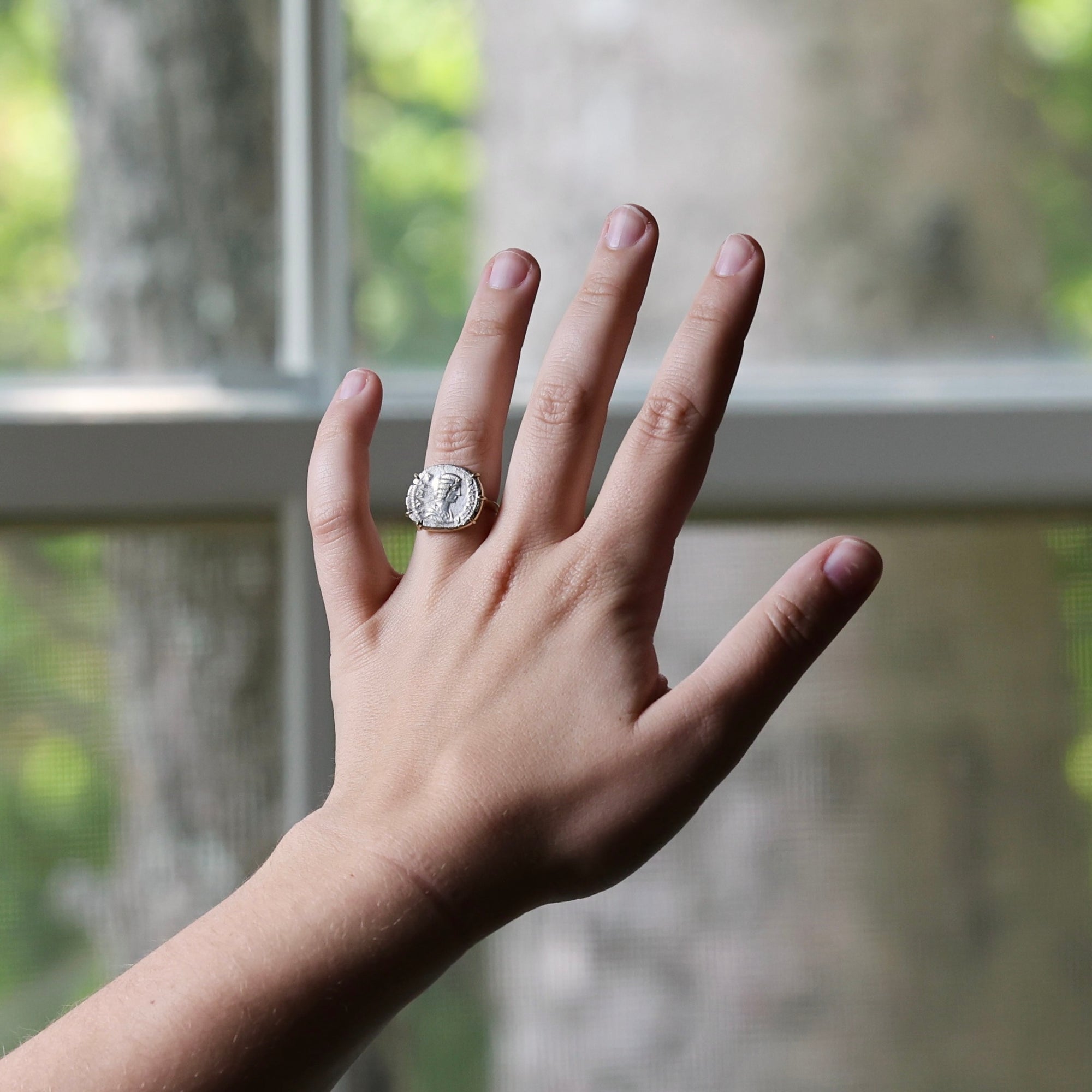 A silver ring with a large, textured circular face is worn on a person's ring finger, with their hand raised in front of a softly blurred window showing green foliage outside.