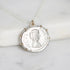A silver pendant necklace shaped like an ancient Roman or Greek coin with a profile portrait, resting on a white marble surface.