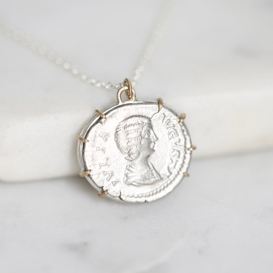 A silver pendant necklace shaped like an ancient Roman or Greek coin with a profile portrait, resting on a white marble surface.