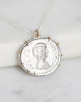A silver pendant necklace shaped like an ancient Roman or Greek coin with a profile portrait, resting on a white marble surface.