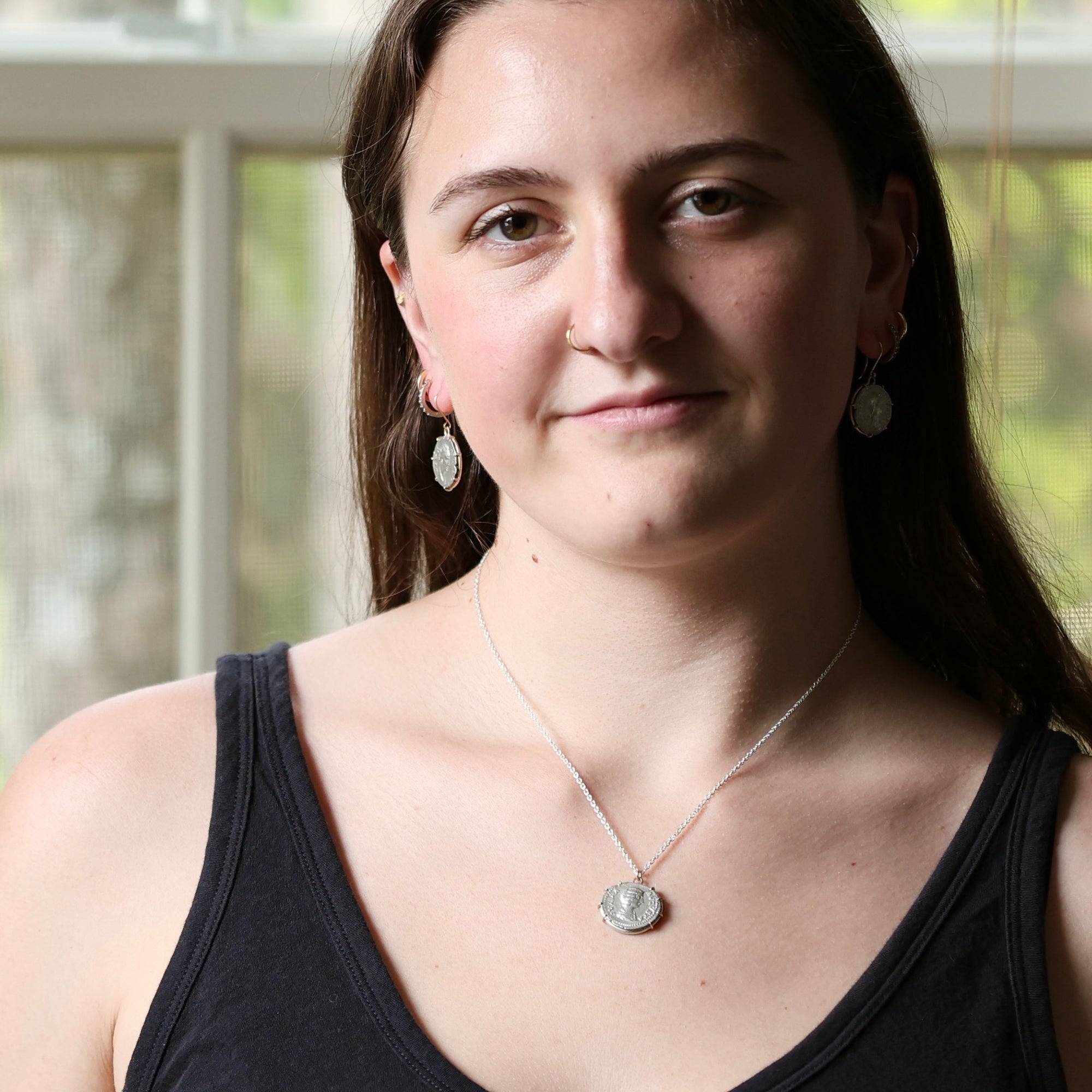 A woman with long brown hair wearing a silver pendant necklace and matching silver earrings, photographed indoors with a softly blurred window background.