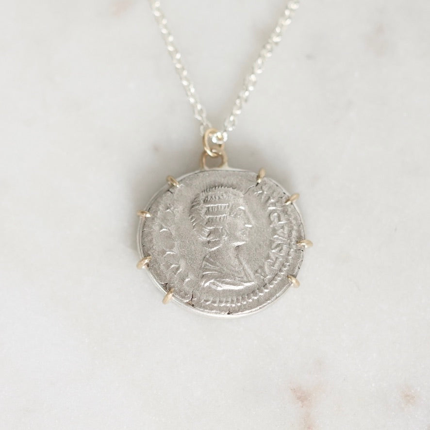A silver pendant necklace featuring an ancient coin pendant with a profile portrait, suspended from a delicate silver chain on a light marble background.