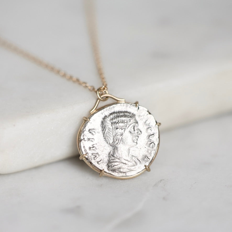 A gold chain necklace with a round silver coin pendant featuring a profile portrait, resting on a white marble block against a light gray marble background.