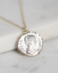 A gold chain necklace with a round silver coin pendant featuring a profile portrait, resting on a white marble block against a light gray marble background.