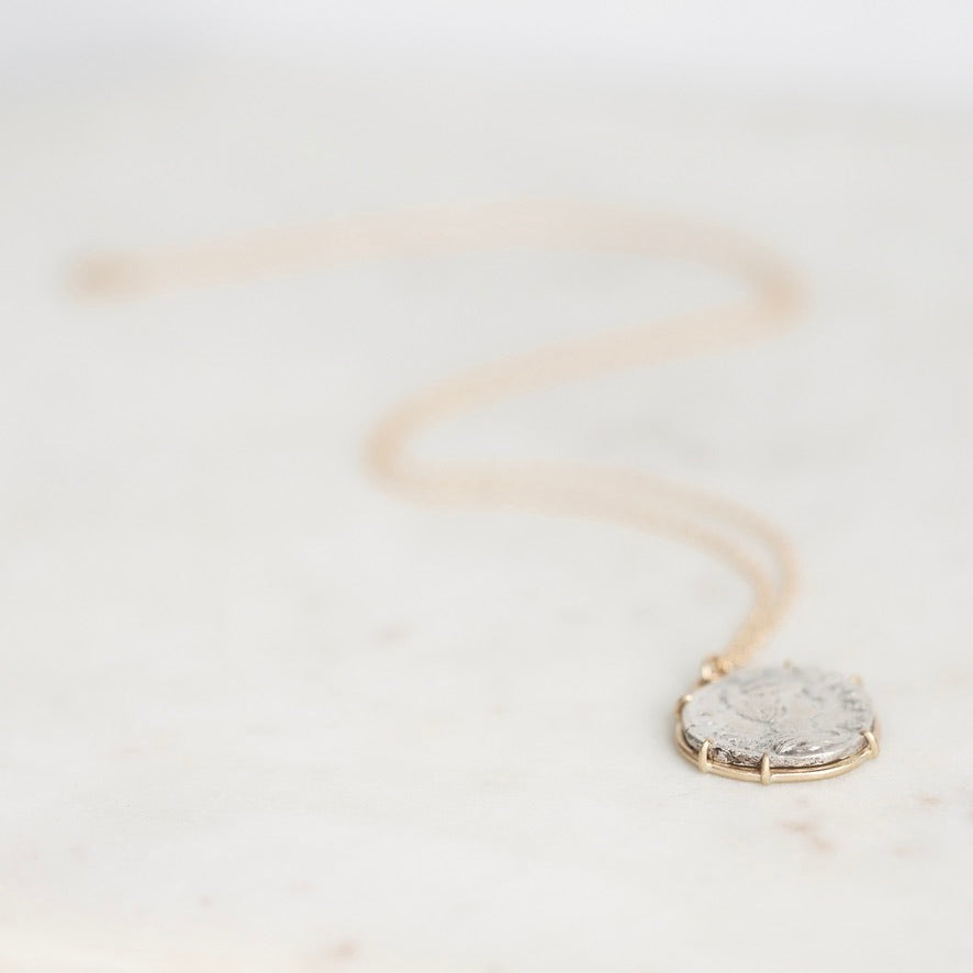 A delicate gold chain necklace with a round, textured silver pendant resting on a light marble surface.