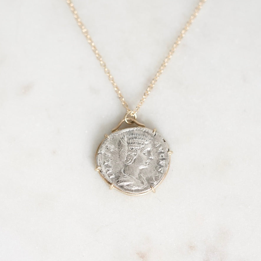 A delicate gold chain necklace featuring a round silver coin pendant with an embossed profile portrait, displayed on a light marble surface.