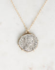 A delicate gold chain necklace featuring a round silver coin pendant with an embossed profile portrait, displayed on a light marble surface.