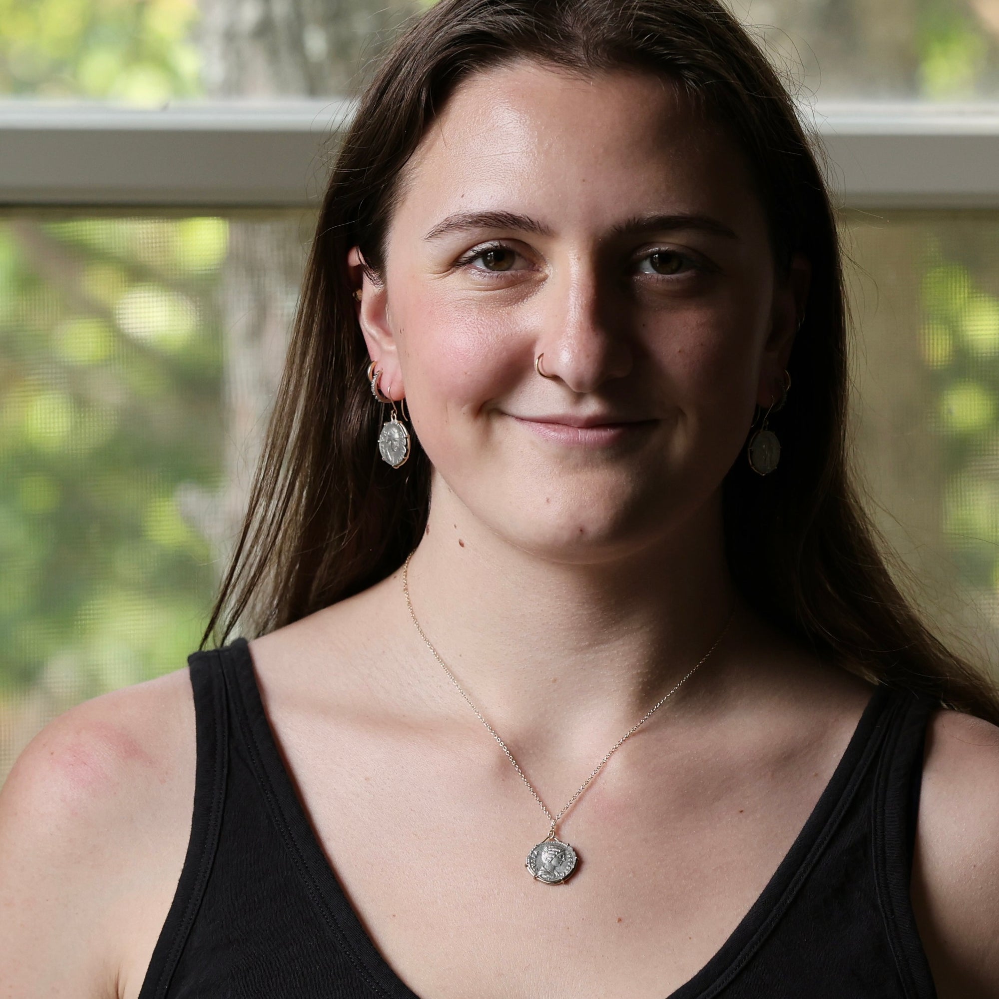 A smiling young woman with long brown hair wearing a black tank top, a silver pendant necklace, and matching silver earrings, photographed indoors with a softly blurred window background.
