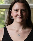 A smiling young woman with long brown hair wearing a black tank top, a silver pendant necklace, and matching silver earrings, photographed indoors with a softly blurred window background.
