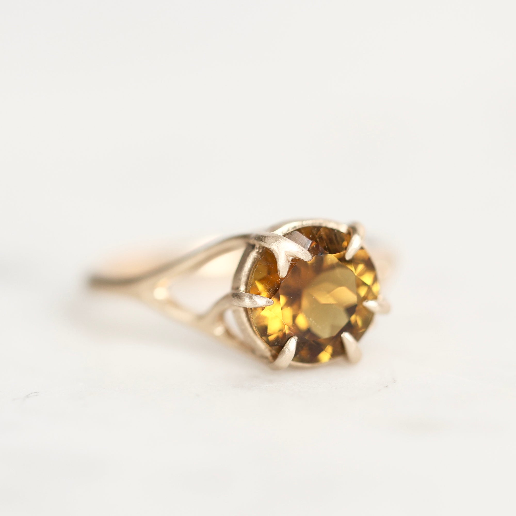 A yellow gold ring with a round-cut gemstone in a prong branch setting, shown on a plain white background.