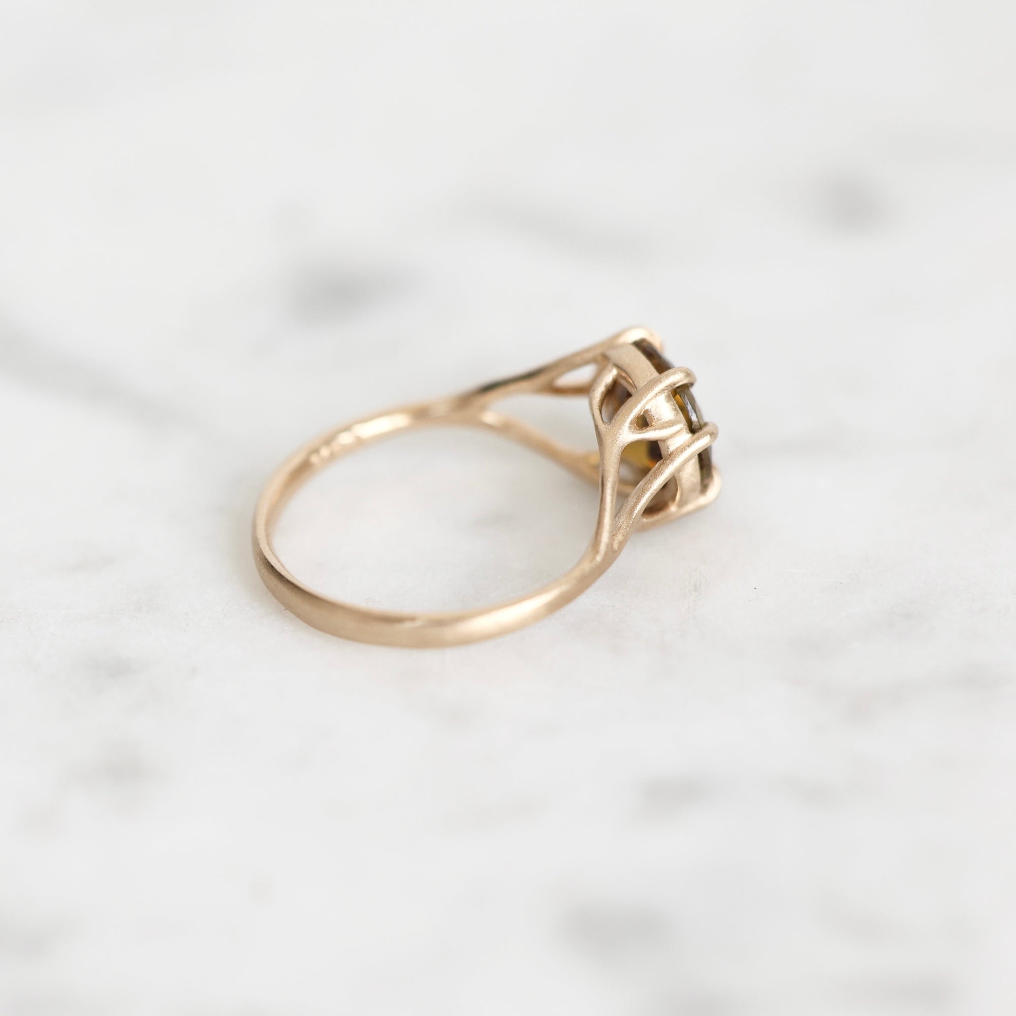 A gold ring with a unique branch band and a small central stone, resting on a light marble surface.