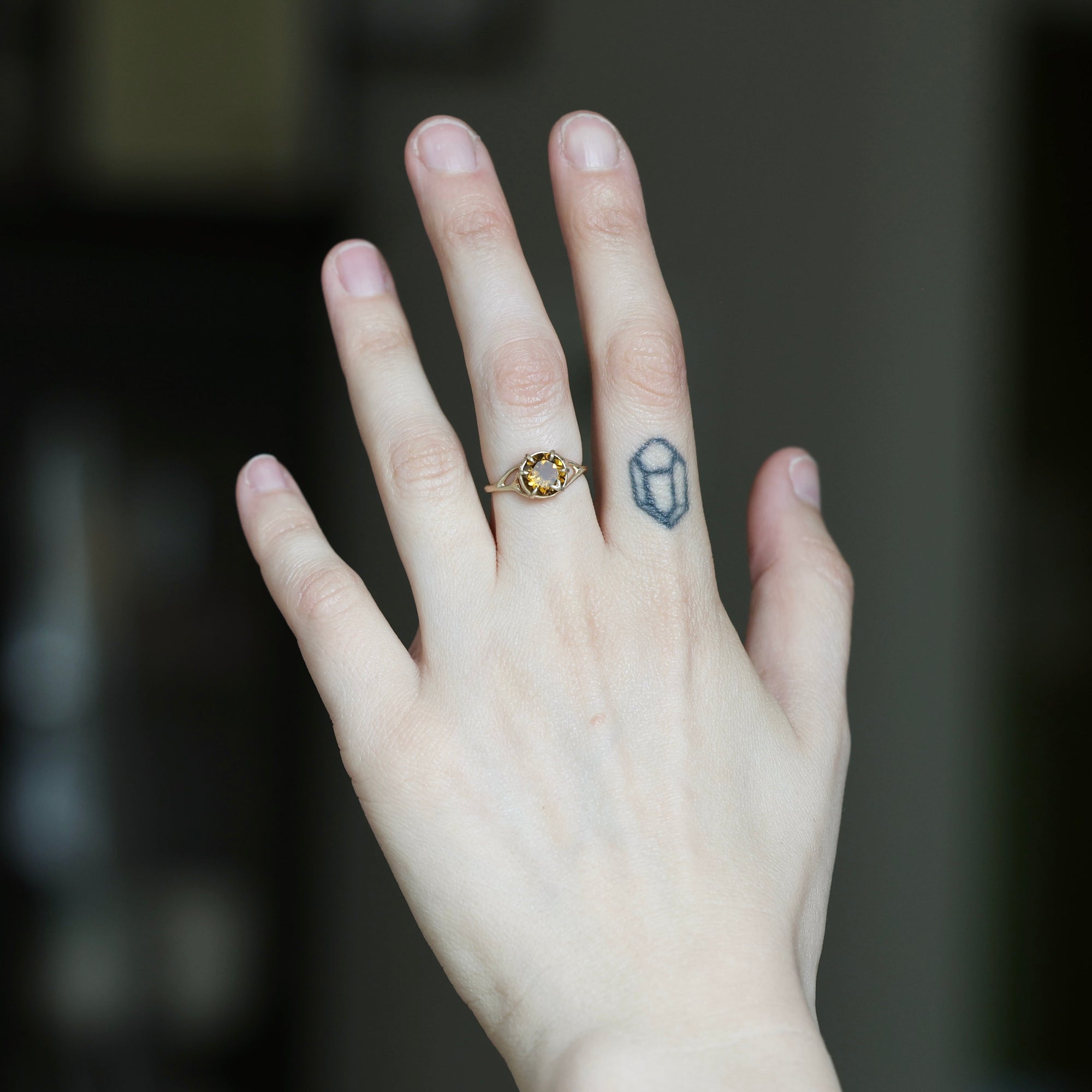 A close-up of a person's hand wearing a delicate gold ring with a small oval yellow gemstone on the ring finger, with a small geometric tattoo on the middle finger, against a dark blurred background.