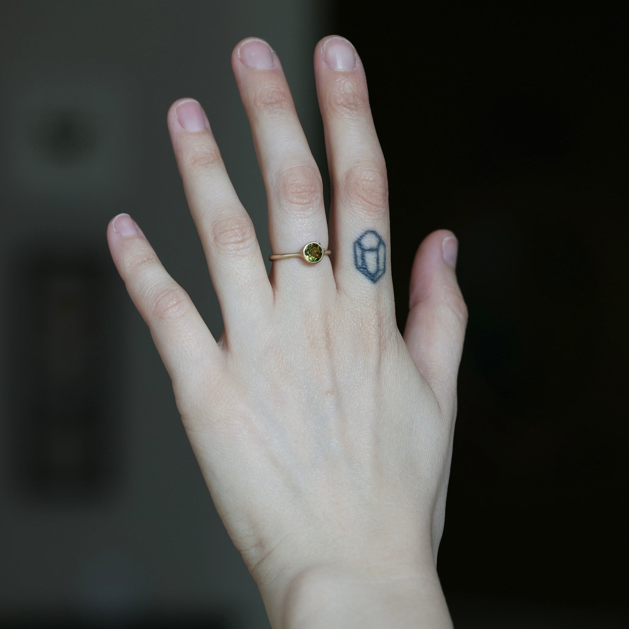 A close-up of a person's hand showing two rings: a delicate gold ring with a small round green gemstone on the ring finger, and a dark blue geometric line-art ring on the middle finger, against a dark blurred background.