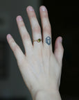 A close-up of a person's hand showing two rings: a delicate gold ring with a small round green gemstone on the ring finger, and a dark blue geometric line-art ring on the middle finger, against a dark blurred background.