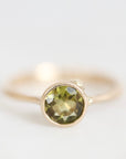 A gold ring with a round faceted olive green gemstone in a prong setting, photographed against a clean white background.