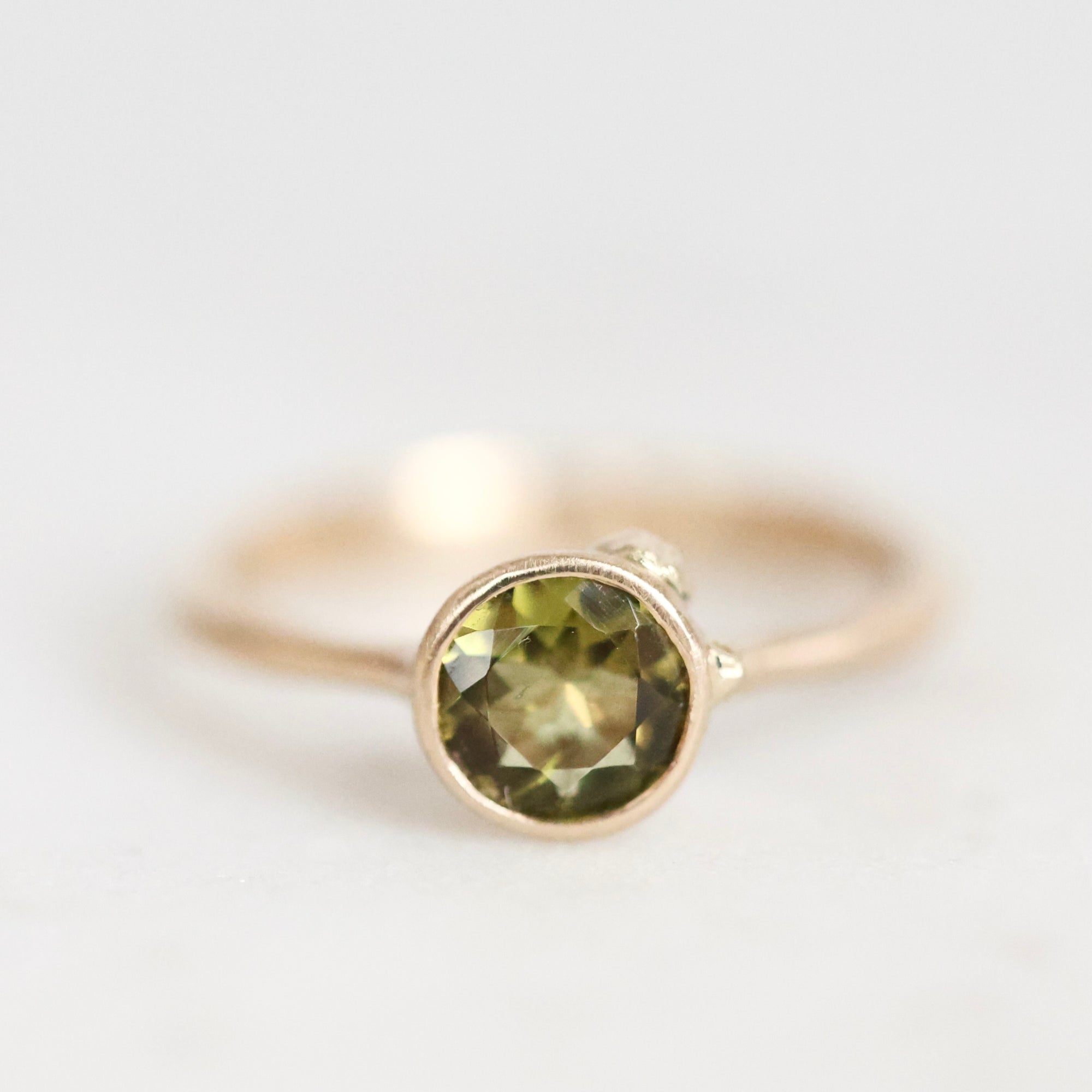 A gold ring featuring a round faceted olive green gemstone in a bezel setting, photographed against a soft, light gray background.