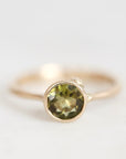 A gold ring featuring a round faceted olive green gemstone in a bezel setting, photographed against a soft, light gray background.