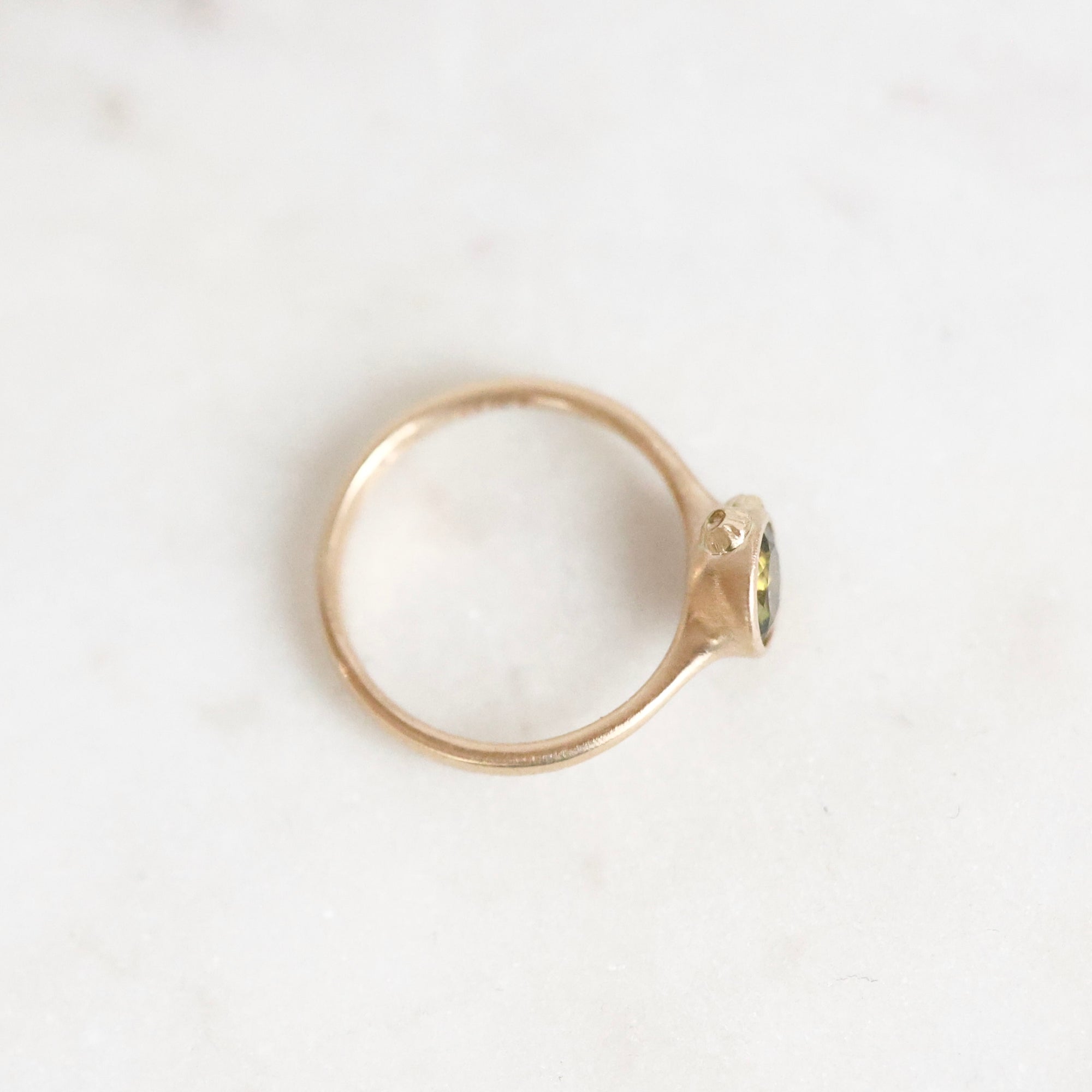 A delicate gold ring with a small oval green gemstone, photographed on a light, textured white surface.