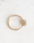 A delicate gold ring with a small oval green gemstone, photographed on a light, textured white surface.