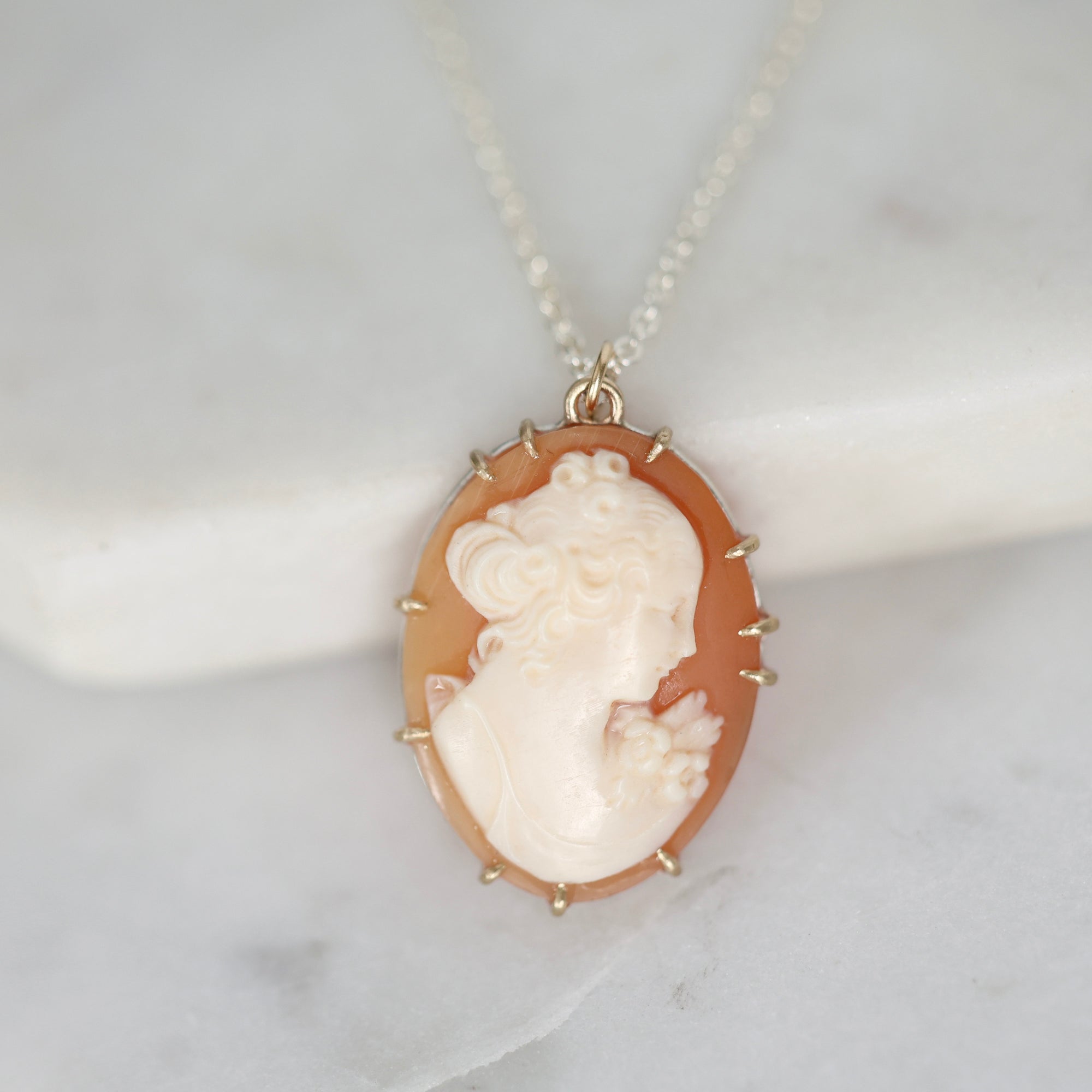 An oval cameo pendant necklace featuring a carved profile of a woman on a peach and cream shell, set in a gold-toned bezel with small spikes, hanging from a silver-toned chain on a white marble surface.