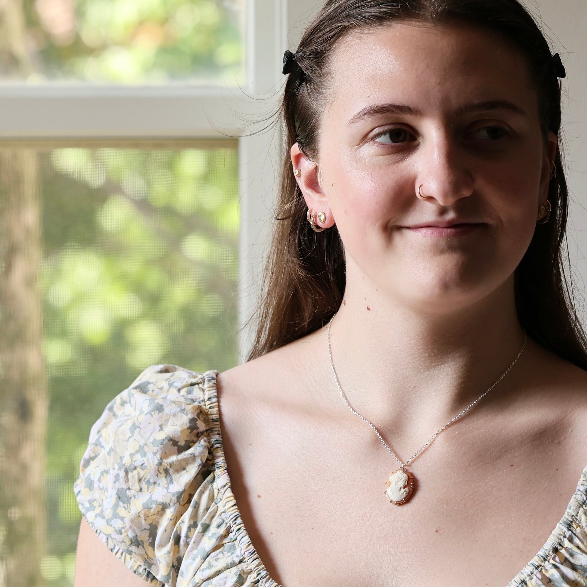 A woman wearing a silver-toned necklace with a small oval shell pendant, photographed indoors near a window with green foliage visible in the background.