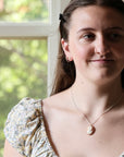 A woman wearing a silver-toned necklace with a small oval shell pendant, photographed indoors near a window with green foliage visible in the background.