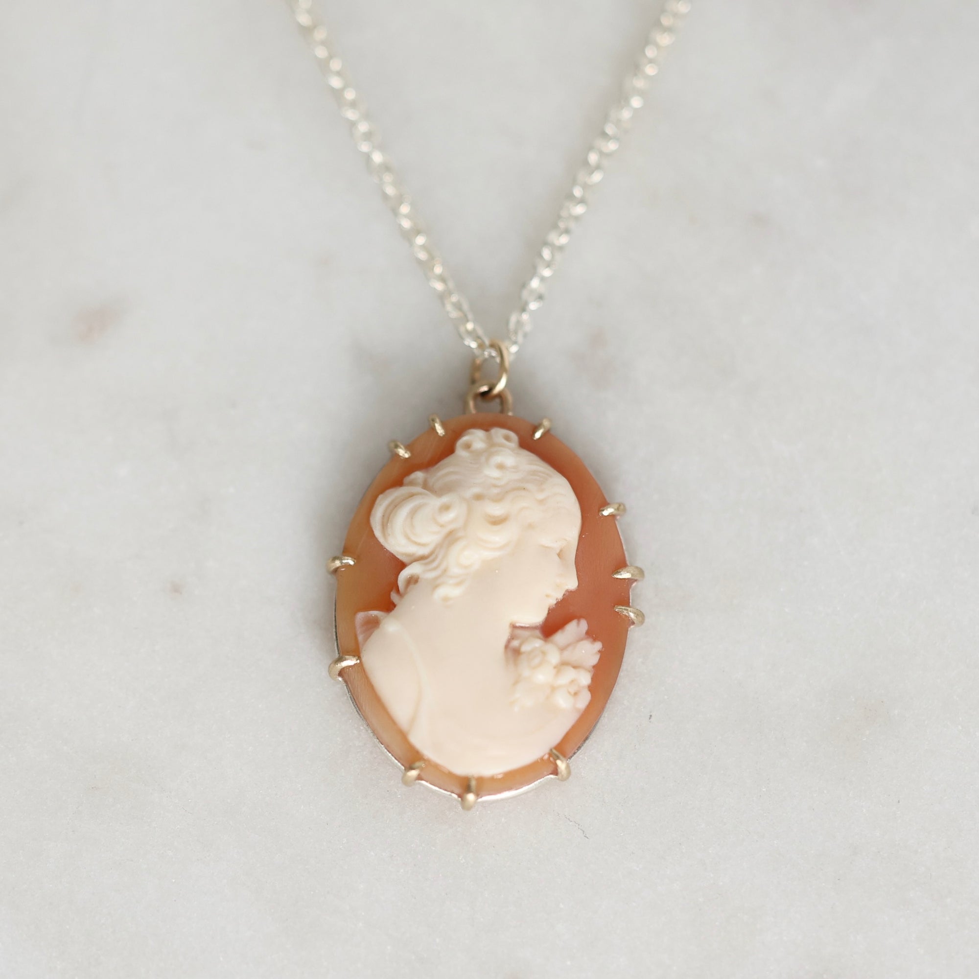 A vintage-style oval pendant necklace featuring a cream-colored carved classical profile on a peach-orange background, set in a gold-toned bezel with small spike details, hanging from a silver-toned chain on a light marble surface.