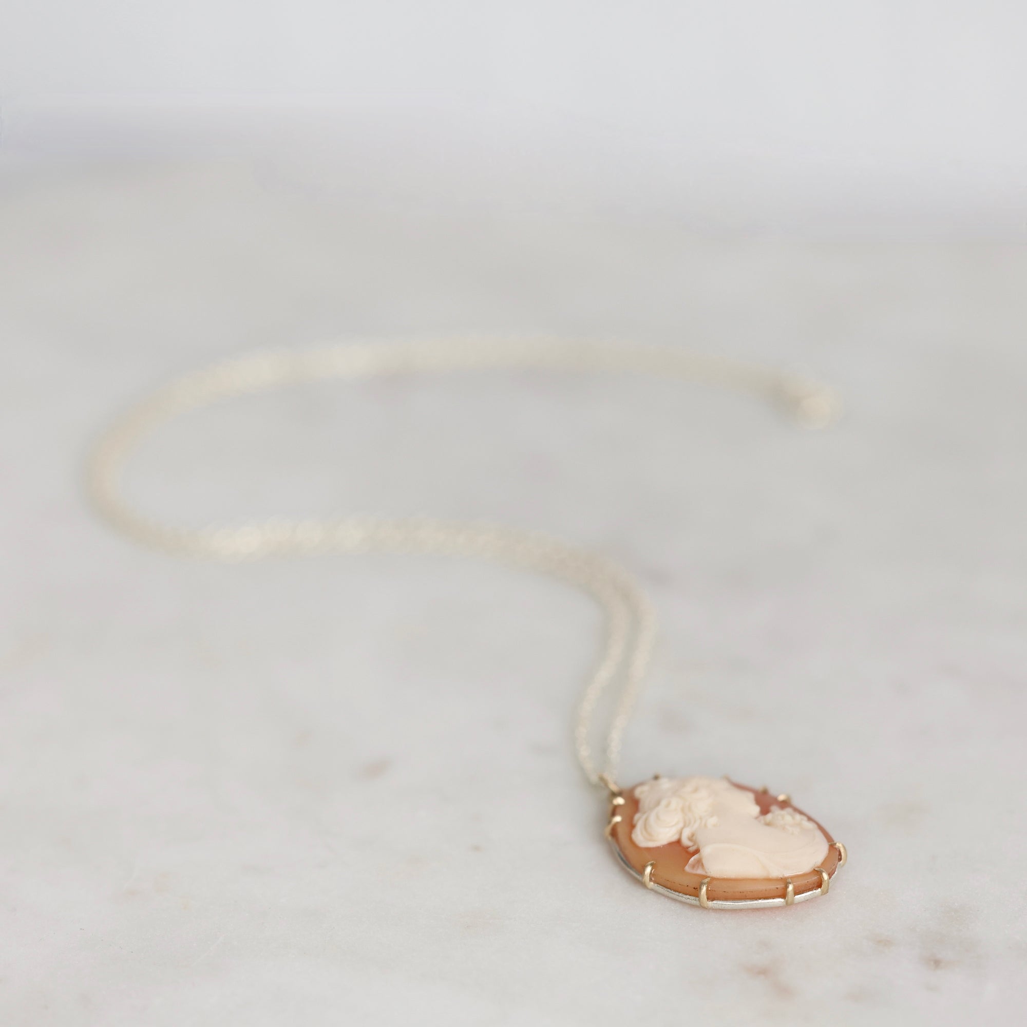 A delicate gold necklace with a cream-colored carved cameo pendant, resting on a light marble surface.