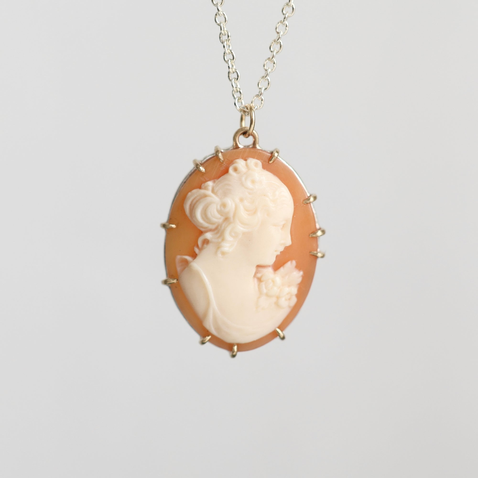 A vintage-style oval pendant necklace featuring a cream-colored carved classical profile on a peach-orange background, set in gold-toned metal with a delicate chain against a light gray background.