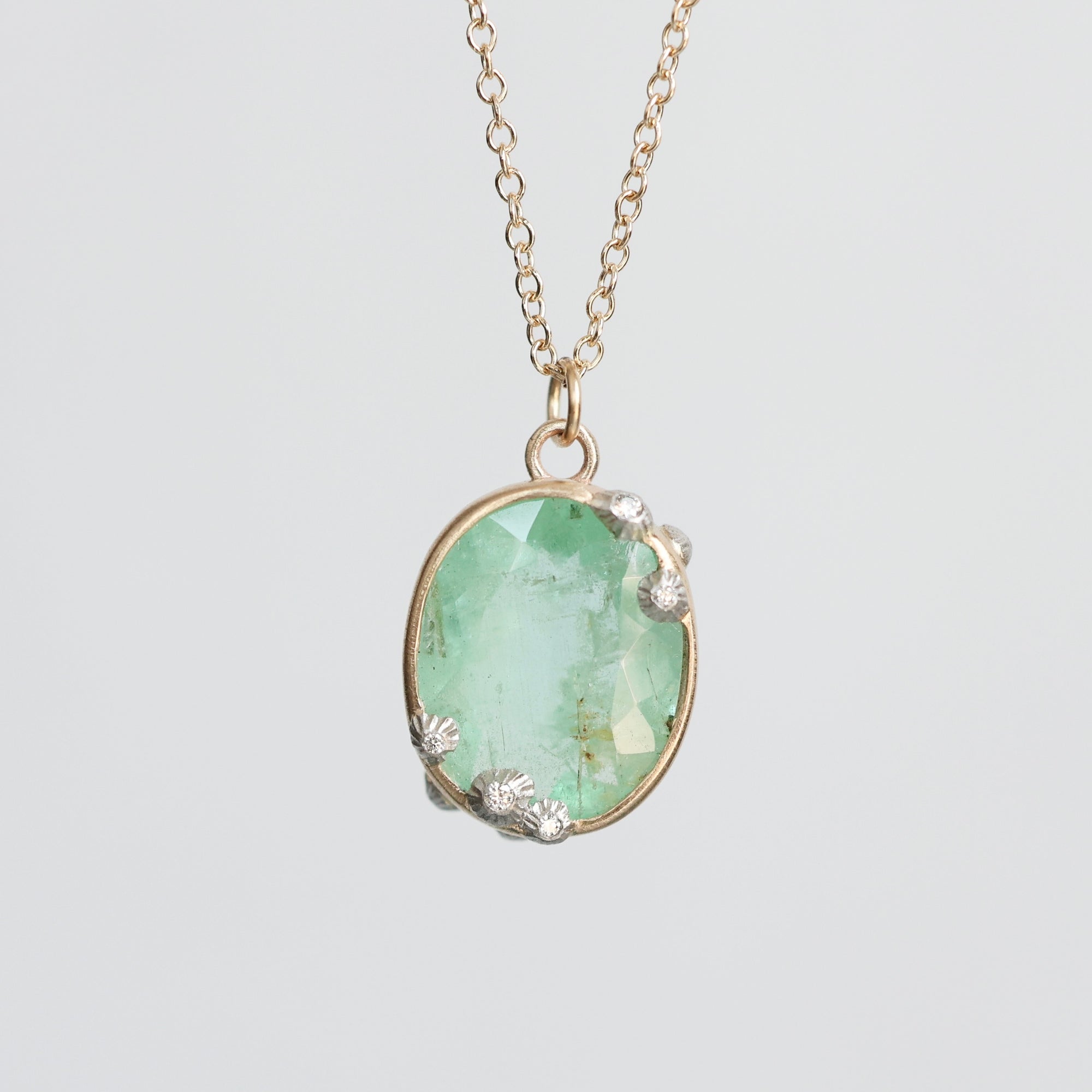 An oval pendant necklace featuring a light green faceted gemstone in a gold-toned setting with textured silver accents, suspended from a delicate gold chain against a light gray background.
